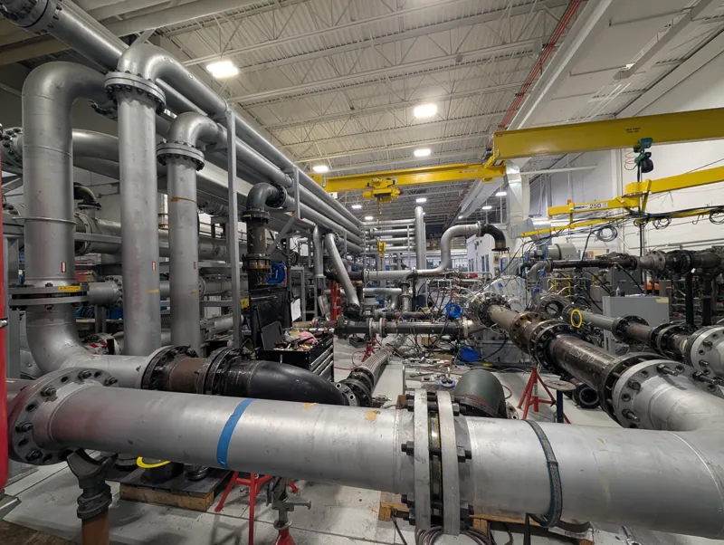 Compressor test bay at Sundyne, Arvada, with large-bore test loop piping, overhead cranes and a centrifugal compressor package on the test bed