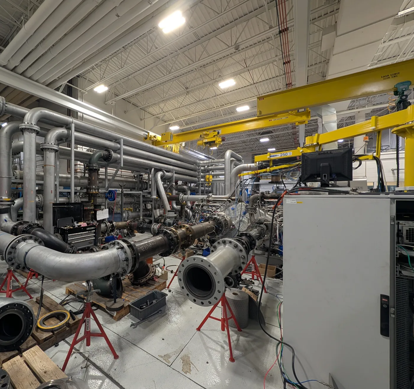 Wide view of the compressor test bay showing test loop piping on pipe stands under overhead cranes